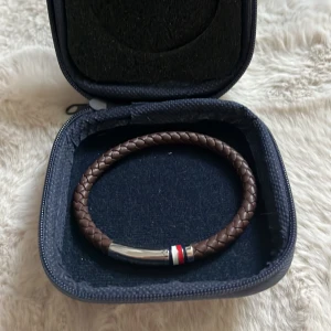 Brunt armband Tommy Hilfiger - Snyggt brunt flätat armband från Tommy Hilfiger med ett stilrent magnetlås i silverfärgad metall. Låset har märkets klassiska ränder i rött, vitt och blått. Kommer i originaletui med logga.