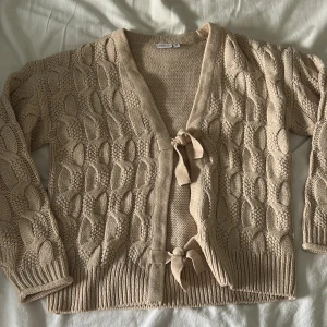 Beige stickad kofta med knyt från Name It - Säljer en beige stickad kofta från Name It med snyggt strukturmönster och två knytband framtill. Koftan har långärmade ärmar med ribbade muddar och v-ringad hals. Jag skulle säga att koftan är lite större i storleken! (Aldrig använt)