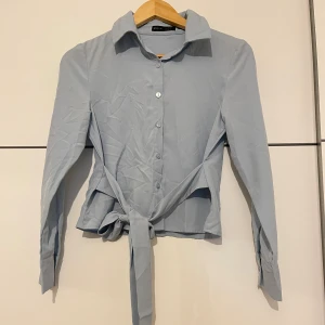 Ljusblå skjorta med knyt  - Snygg ljusblå skjorta  med klassisk krage, långa ärmar och knappar framtill. Skjortan har långa band i midjan som kan knytas för en trendig look. Perfekt till jeans eller kjol för en stilren vibe.