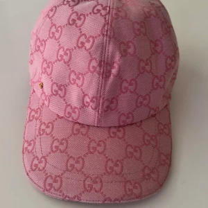Rosa Gucci keps med GG-mönster - Snygg rosa keps från Gucci med ikoniskt GG-mönster över hela kepsen. Kepsen har böjd skärm, justerbar rem i rosa läder och en liten guldfärgad detalj på sidan. Insidan är ljusbeige med Gucci-etikett. Perfekt för dig som vill sticka ut med lyxig streetstyle.