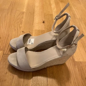 Kilklacksko - Helt oanvända. Snygga beige sandaler med kilklack och flätad sula. Skorna har öppen tå, bred rem över foten och justerbar ankelrem med spänne. Ovandelen är i mockaimitation och sulan har ett repinspirerat mönster. Perfekta för sommarens alla outfits. 