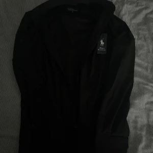 Svart skjorta Polo Ralph Lauren M - Svart vindjacka från Polo Ralph Lauren i slim fit-modell. Jackan är tillverkad i 100% bomull och har klassisk krage, knäppning framtill och broderad logga på bröstet. Perfekt för dig som gillar stilren och enkel design.