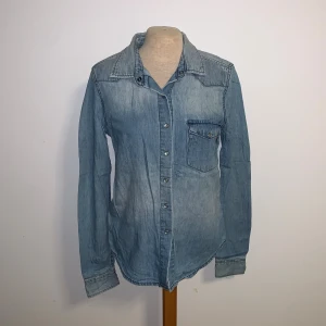 Ljusblå jeansskjorta från Crocker - Snygg ljusblå jeansskjorta från Crocker i klassisk modell med bröstficka och knappar framtill. Skjortan har lång ärm, perfekt att bära öppen eller stängd för en cool look.