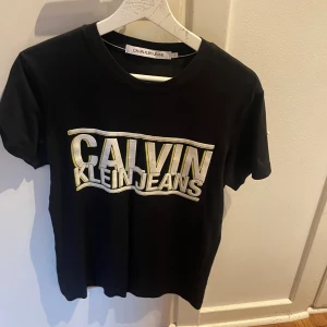 Svart Calvin Klein Jeans t-shirt - Svart t-shirt från Calvin Klein Jeans med stort, glansigt logotryck i silver och guld på bröstet. Klassisk rund hals och korta ärmar. Perfekt för dig som gillar stilrena och trendiga märkesplagg med lite extra attityd.