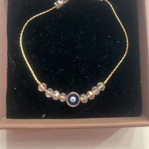 Guldigt armband med blått öga - Snyggt armband med tunn guldfärgad kedja och transparenta pärlor. I mitten sitter en blå evil eye-berlock som ger en cool och mystisk vibe. Perfekt accessoar för att lyfta din stil.