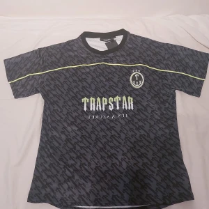 Trapstar svart fotbollströja S/M - Svart och grå fotbollströja från Trapstar med korta ärmar och grafiskt mönster. Tryck med Trapstar-logga och texten 'IT'S A SECRET' på bröstet samt siffran 22 på ryggen. Neongul detalj över bröst och rygg. Tillverkad i lätt och ventilerande material. Perfekt för träning samt daglig användning.