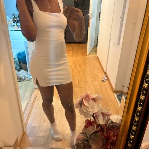 Vit tight kortklänning med slits - Supersnygg vit kortklänning med breda axelband och liten slits på ena sidan. Klänningen har en tight passform och fyrkantig ringning, perfekt för dig.