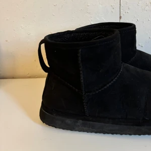 Svarta boots i mocka - Svarta boots i mjuk mocka med rund tå och platt sula. Skorna har en klassisk, enkel design med kort skaft och dragflik baktill. Perfekta för kyliga dagar och passar till många olika outfits.