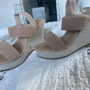 Beige kilklackade sandaler med remmar - Snygga sandaler med beige textilremmar och hög kilklack i flätad look. Skorna har öppen tå och breda band över foten för en stabil känsla. Perfekta för sommardagar när du vill ha lite extra höjd och stil.