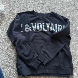 Svart sweatshirt från Zadig & Voltaire - Cool svart sweatshirt från Zadig & Voltaire med stort silvrigt tryck framtill. Tröjan har långa ärmar och en snygg dragkedja i sidan som detalj. Perfekt för dig som gillar streetstyle och vill ha något som sticker ut lite extra.