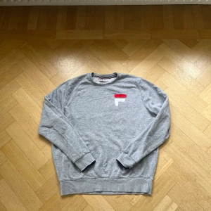 Grå sweatshirt från FILA i bomull Storlek M - Grå crewneck sweatshirt från FILA med broderad logga i rött och vitt på bröstet. Tröjan har långa ärmar, ribbade muddar och klassisk passform. Tillverkad i mjuk bomullsmix, perfekt för en chill och sportig stil.