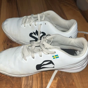 Vita fotbollsskor från Salming strl 36 - Säljer ett par vita Salming fotbollsskor i storlek 36. Skorna har snörning, svart logga på sidan och en liten svensk flagga. Ovandelen är i syntetmaterial och sulan har hexagonmönster för bra grepp på planen.