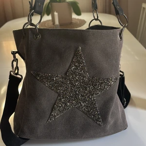 Grå axelväska med glittrig stjärna - Cool grå axelväska med en stor glittrig stjärna framtill. Väskan har bred svart axelrem. 