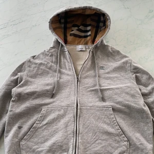 Grå hoodie från Burberry med dragkedja - Snygg grå hoodie från Burberry med klassiskt rutigt foder i huvan och broderad logga på bröstet. Tröjan har dragkedja, snörning i huvan och stora fickor framtill. Perfekt för dig som gillar stilren design med lyxig touch. Den är i nyskick och inga konstigheter finns på tröjan, allt funkar som det ska👍pris kan även diskuteras !