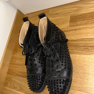 Christian Louboutin svarta sneakers med nitar - Svarta high-top sneakers i skinn från Christian Louboutin med ikonisk röd sula och logga på sidan. Skorna är täckta av svarta nitar för en edgy look och har snörning framtill. Perfekta för dig som vill sticka ut med lyxig streetstyle.