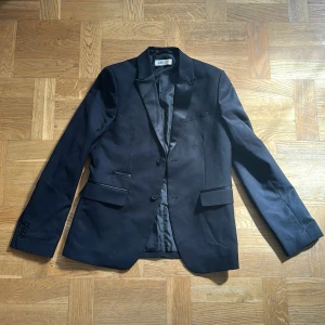 Svart kavaj från H&M, storlek 158 (S) - Snygg svart kavaj från H&M i storlek 158. Klassisk modell med två knappar framtill, smala slag och fickor med lock. Kavajen har en stilren look och är fodrad med slät insida. Perfekt för dig som vill ha en clean och dressad stil.