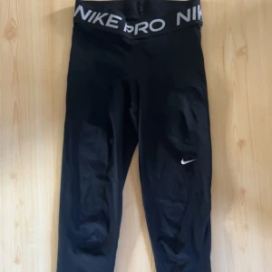 Nike Pro svarta leggings - Svarta Nike Pro leggings med bred resår i midjan och vit logga. Tillverkade i Dri-FIT-material som andas och transporterar bort fukt. Sitter tight och är perfekta för träning eller sport. Snygg och sportig look med diskret Nike Swoosh på benet. Den har en liten skada uppe vid midjan men knappt något man ser. Byxorna är o storlek S men är väldigt liten i storlek. (OBS! på bilden ser man inte hela byxorna så fråga om mer bilder om ni behöver)