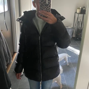 Massimo dutti jacka - Säljer en svart pufferjacka med stor huva och dragkedja framtill. Jackan är vadderad och har en lite längre modell som håller dig varm. Perfekt för kalla dagar och riktigt snygg till jeans eller mjukisbyxor.