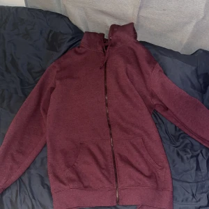 Vinröd hoodie med dragkedja - Vinröd hoodie med hel dragkedja och huva. Tröjan har långa ärmar, känguruficka framtill och snörning i huvan. Perfekt för en chill och avslappnad stil.