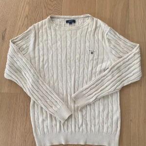 Beige kabelstickad tröja från GANT - Kabelstickad beige tröja från GANT med rund halsringning och diskret broderad logga på bröstet. Tröjan har långa ärmar och ribbade muddar vid ärmslut och nederkant. Perfekt för lager-på-lager och snygg till jeans.