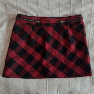 Rutig röd/svart kort kjol med dragkedjsdetaljer - storlek 40 - Snygg kortkjol i röd och svart rutig design med dragkedsdetaljer i sidan. Kjolen har en klassisk A-linjeform och är perfekt för dig som gillar en edgy stil. Materialet är lite tjockare, liknande filt/ylle, och med ett glansigt fodertyg. Storlek 40. 