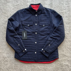 Vändbar quiltad jacka från J.Lindeberg - Snygg vändbar jacka från J.Lindeberg med ena sidan i marinblått och andra i rött med svart detalj på ryggen. Quiltad design, två bröstfickor med knappar och klassisk krage. Perfekt för dig som vill variera din stil och hålla värmen. Säljer pågrund av att den bara hängt i garderoben. 