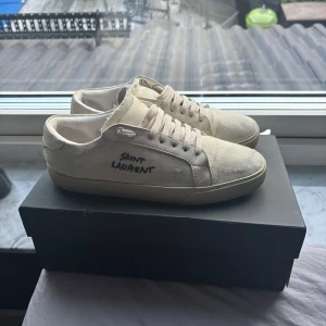 Saint Laurent sneakers - Snygga vita sneakers från Saint Laurent i canvas med broderad logga på sidan. Skorna har rund tå, platt sula och klassisk snörning. Perfekta för dig som gillar stilren och minimalistisk design med exklusiv känsla. Riktigt bra skick 9-10! Pris kan  diskuteras vid snabb affär. Äktihetsbevis finns🙌