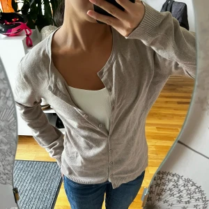 Beige kofta med knappar - Säljer en beige kofta med rund hals och knappar hela vägen framtill. Koftan har långa ärmar med ribbade muddar och är gjord i mjukt material som känns skönt mot huden. Perfekt att slänga över en topp och matcha med jeans för en chill look.