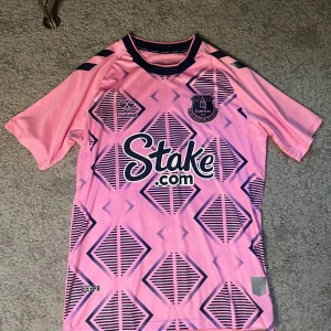 Everton fotbollströja  - Säljer en Everton fotbollströja från Hummel i ljusrosa med mörkblå geometriskt mönster på framsidan. Kortärmad med rund hals och tryckta detaljer, inklusive klubbmärke och sponsor. Tillverkad i lätt polyester som andas, perfekt för träning eller match.
