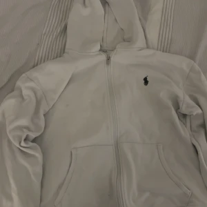 Vit hoodie från Polo Ralph Lauren - Vit hoodie från Polo Ralph Lauren med dragkedja och klassisk broderad logga på bröstet. Tröjan har huva, kängurufickor och är tillverkad i mjuk bomullsmix. Perfekt för en chill och stilren look.