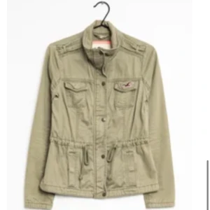 Trendig jacka från Hollister  - Jättesnygg trendig olivgrön jacka från Hollister med dragkedja och knappar framtill. Jackan har fyra fickor, snörning i midjan och klassisk krage. Perfekt för vår och höst. Liten logga på bröstfickan ger extra stil.