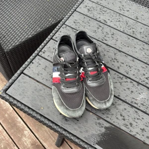 Moncler sneakers med färgdetaljer - Snygga svarta Moncler sneakers med detaljer i rött, vitt och blått på sidan. Skorna har svarta snören, rund tå och en beige sula. Ovandelen är i en mix av skinn och textil, med Moncler-logga på plösen. Perfekta för dig som gillar stilrena men ändå unika sneakers. Köpte i moncler butik 2023! Nypris 5300