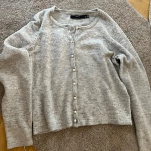 Grå kofta med pärlknappar Vero Moda - Superfin ljusgrå kofta från Vero Moda med långa ärmar och snygga pärlknappar framtill. Mjuk och skön, perfekt att slänga på sig över en topp. Enkel och stilren design som funkar till det mesta.
