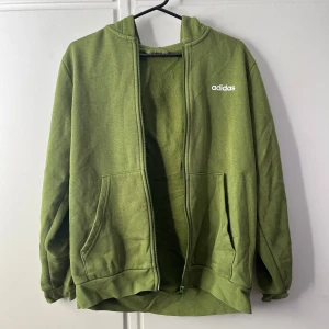 Grön Adidas hoodie med dragkedja - Säljer en grön hoodie från Adidas med klassiska vita ränder längs ärmarna och vit Adidas-logga på bröstet. Tröjan har huva, dragkedja framtill och två stora fickor. Perfekt för en sportig och chill stil.