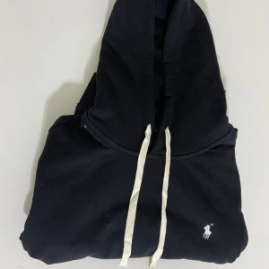 Svart hoodie från Polo Ralph Lauren - Svart hoodie från Polo Ralph Lauren med vit broderad logga på bröstet och vita snören i huvan. Klassisk känguruficka framtill och ribbade muddar vid ärmslut och nederkant. Perfekt för en chill och stilren look.