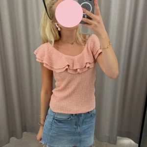 Ljusrosa volangtopp från Lindex - Supersöt ljusrosa topp från Lindex med volangdetaljer längs den vida urringningen. Toppen är ärmlös och har en strukturerad, lite krinklad bomullskänsla. Perfekt till jeanskjol eller shorts för en fräsch sommarlook!