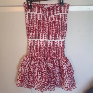 Rödvit klänning från design by amor - Aldrig använd!!! Supersöt kortklänning från Designby Amor i storlek S. Klänningen är axelbandslös med volangig kjol och har ett broderat prickigt mönster i rött på vit botten. Materialet är bomull och ger en luftig känsla, perfekt för varma dagar. Smockad överdel för en tight passform.