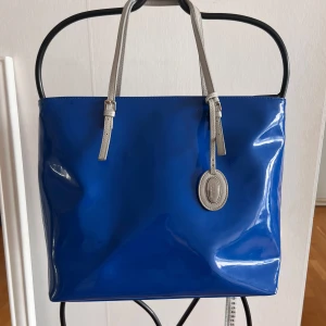 Blå shoppingväska från Furla - Snygg blå shoppingväska från Furla i glansigt syntetmaterial med gråa skinnhandtag. Väskan har Furla-logga på en tag och stängs med dragkedja. Rymlig insida med innerfack och stilren design som sticker ut.