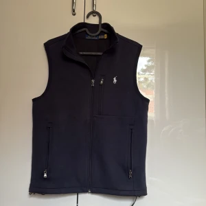 Mörblå väst från Polo Ralph Lauren XS - Stilren mörkblå väst från Polo Ralph Lauren i storlek XS. Västen har hög krage, tre fickor med dragkedja och klassisk logga broderad på bröstet. Perfekt för lager-på-lager och enkel att matcha med olika outfits. Slutsålt nästan överallt och kostar 2200kr ny. Mycket bra skick