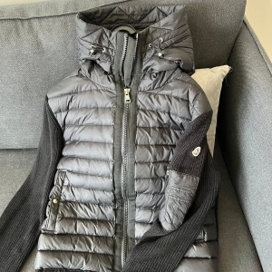 Svart Moncler pufferjacka med stickade ärmar - Snygg svart pufferjacka från Moncler med stickade ärmar och huva. Jackan har quiltad framsida i glansig polyester, dragkedja framtill och praktiska fickor. Ärmarna är ribbstickade och har Moncler-logga på vänster arm. Perfekt för kyliga dagar.