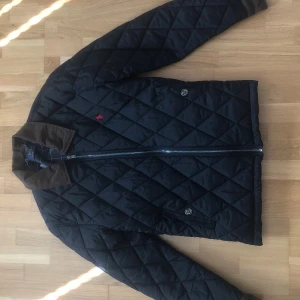 Svart quiltad jacka Polo Ralph Lauren - Quiltad svart jacka från Polo Ralph Lauren med brunt manchesterkrage, dragkedja framtill och broderad röd logga på bröstet. Jackan har lång ärm och klassisk passform, perfekt för höst och vår. Snygg detalj med rutigt innerfoder.