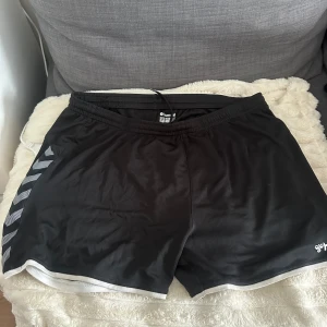 Svarta sportshorts från Hummel XL - Svarta sportshorts från Hummel i storlek XL. De har elastisk midja, vita detaljer längs bensluten och klassiska Hummel-pilar i vitt på sidorna. Perfekta för träning eller sport. Tillverkade i lätt och snabbtorkande polyester perfekt till träning 