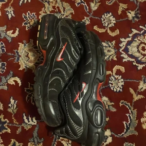 Nike Air Max Plus svarta sneakers - Nike Air Max Plus sneakers i svart med röda och vita detaljer. Snygg vågig design på ovandelen, synlig Air Max-dämpning och coola detaljer på sulan. Skorna har snörning och en sportig vibe, perfekta för dig som gillar streetwear och unika sneakers.