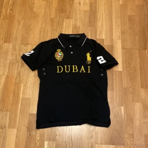 Cheif keif polo Svart Polo Ralph Lauren Dubai t-shirt - Svart t-shirt från Polo Ralph Lauren med gul broderad Dubai-text och emblem på bröstet. Klassisk polokrage med vita detaljer, korta ärmar med siffran 2 i vitt på både ärm och rygg. Materialet är mjuk bomull och passformen är normal. Perfekt för dig som gillar sportig och exklusiv stil.