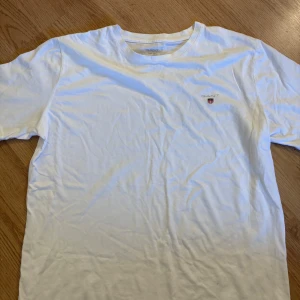 Vit t-shirt från GANT med logga - Vit t-shirt från GANT i mjuk bomull. Klassisk rund hals och korta ärmar. På bröstet finns en liten GANT-logga med sköld i rött, blått och vitt. Enkel och clean design som funkar till jeans eller shorts.