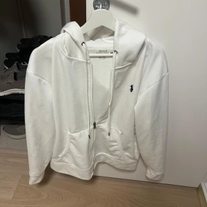 Vit hoodie från Polo Ralph Lauren - Snygg vit hoodie från Polo Ralph Lauren med dragkedja, huva och klassisk logga broderad på bröstet. Tröjan har känguruficka och justerbara snören i huvan. Perfekt för en chill och stilren look. Enda defekten är att snörena är lite långa, har bytat ut orginalsnörena.