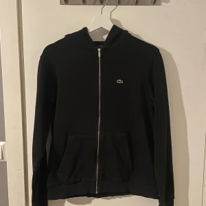 Lacoste zip hoodie - Svart hoodie från Lacoste med klassisk krokodillogga på bröstet. Tröjan har huva, hel dragkedja framtill. Perfekt för dig som gillar stilren och sportig look. Materialet känns mjukt och bekvämt. Storleken är 176 och passar som en S. Pris kan självklart diskuteras