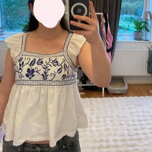 Vit blus med blå broderi och volang - Supersöt vit blus med blått broderat blommönster framtill och volangärmar. Finns några fläckar invändigt men inget som syns, blid kan skickas vid intresse💗