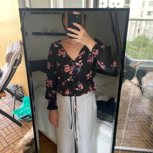 Svart blommig blus från H&M Divided - Säljer en svart blus från H&M Divided med röda och rosa blommor. Blusen har v-ringning, snörning framtill och långa ärmar med smock vid ärmslut. Croppad passform och lätt, flowigt material som känns skönt mot huden. Perfekt för dig som gillar romantiska detaljer.