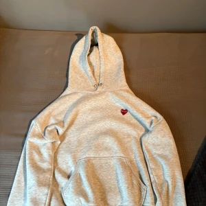 Grå hoodie Comme des Garçons Play - Grå hoodie från Comme des Garçons Play med klassisk huva och magficka. På bröstet sitter det ikoniska röda hjärtat med ögon. Perfekt för dig som gillar streetwear och enkel stil. OBS inte original
